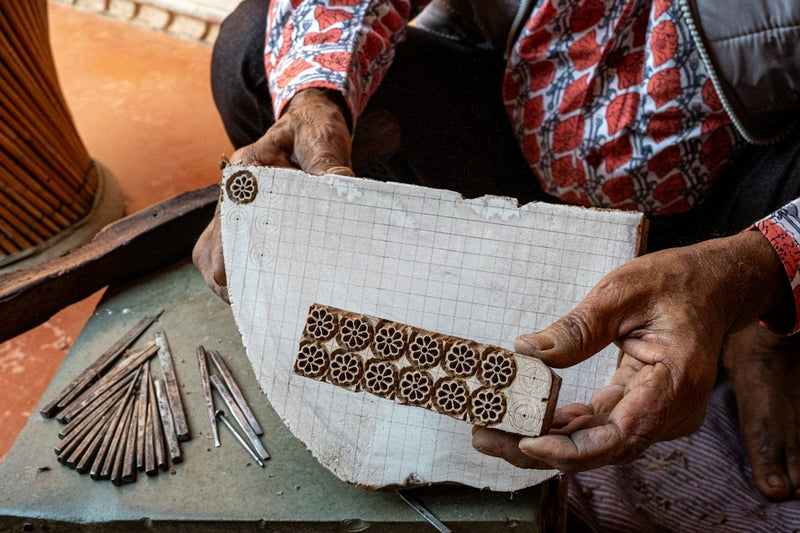 Artisan in Jaipur, Rajasthan holding a carved wooden printing block and design sketch, with carving tools nearby, evoking the hand block printing tradition behind Sanganer and Bagru prints