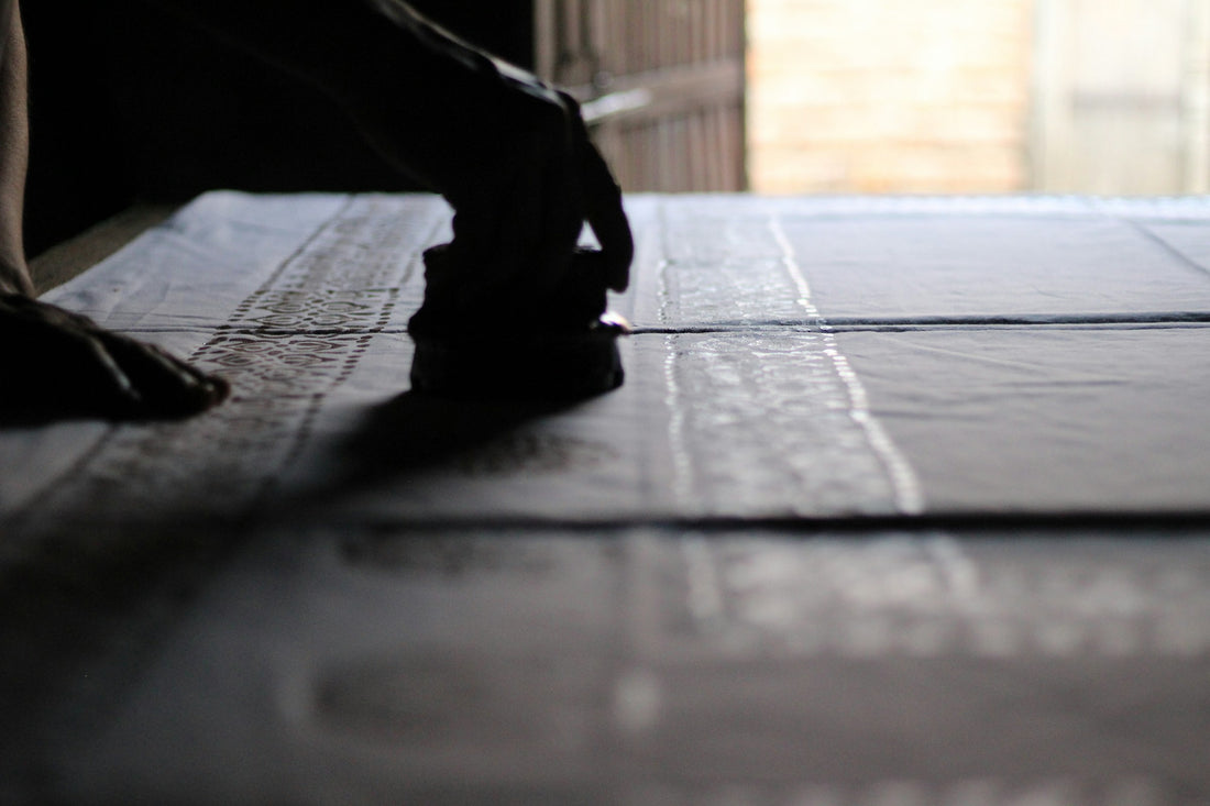 Hand pressing a carved wooden block onto indigo-blue printed fabric in a rural Indian village, evoking Rajasthan's dabu and block printing tradition