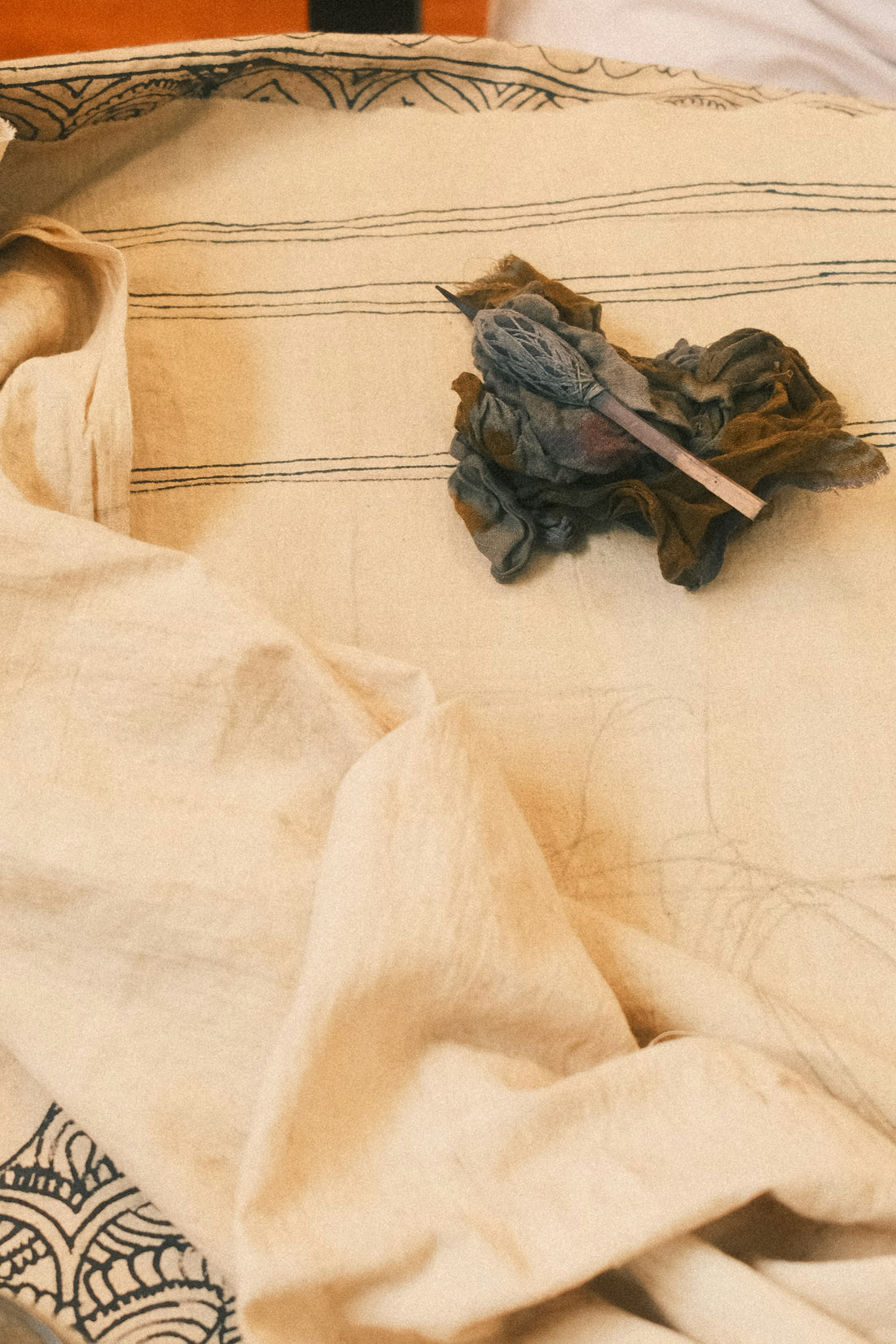 Bundled block-printed fabric scraps and a carved wooden printing block, evoking the hand block printing and natural dyeing tradition behind Ajrakh