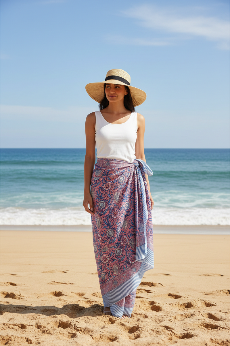 Woman on a beach wearing a white top and patterned sarong with a straw hat.