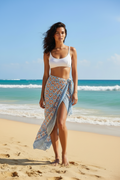 Woman standing on a beach wearing a white top and patterned sarong with blue ocean and sky in the background.