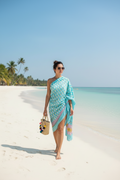 Woman in a blue patterned dress standing on a sandy beach with clear blue water and palm trees in the background.
