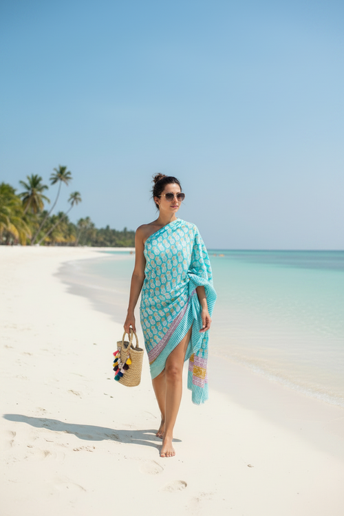 Woman in a blue patterned dress standing on a sandy beach with clear blue water and palm trees in the background.