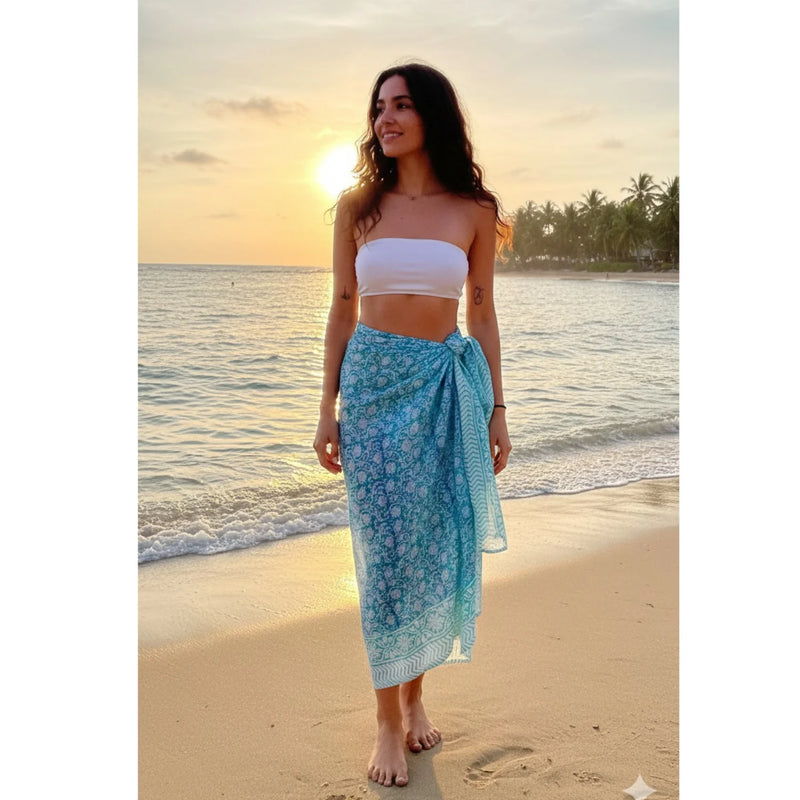 Woman in a white top and blue floral skirt standing on a beach at sunset.